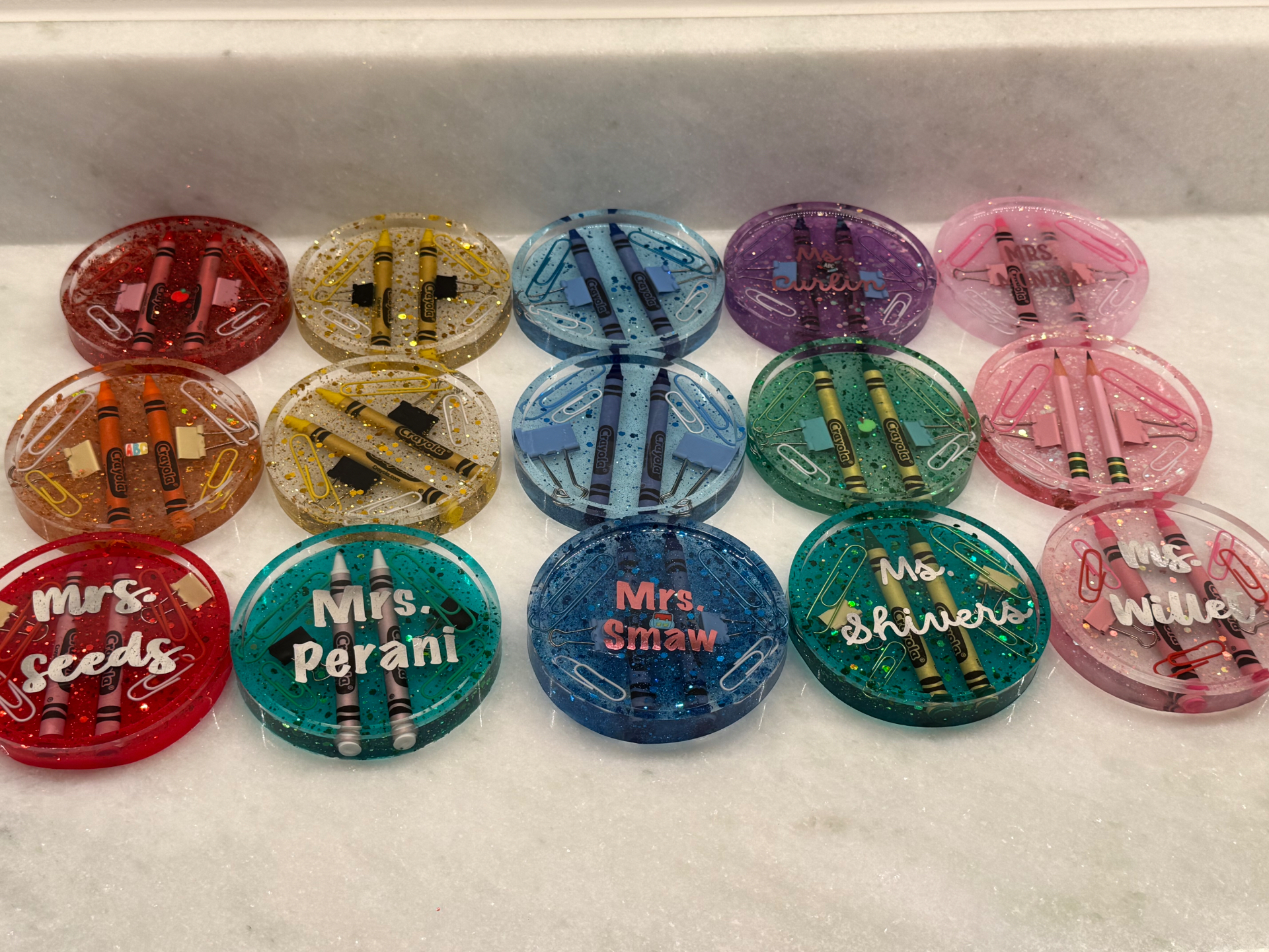 Collection of colorful personalized resin coasters with teacher names, crayons, paperclips, and school-supply inclusions